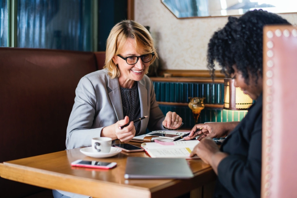 Two ladies out in a cafe sitting at a booth having a meeting with paperwork, coffee and phones on the table. One lady is looking down at the paperwork in front of her while the other lady is looking happily at the paperwork across the table.
