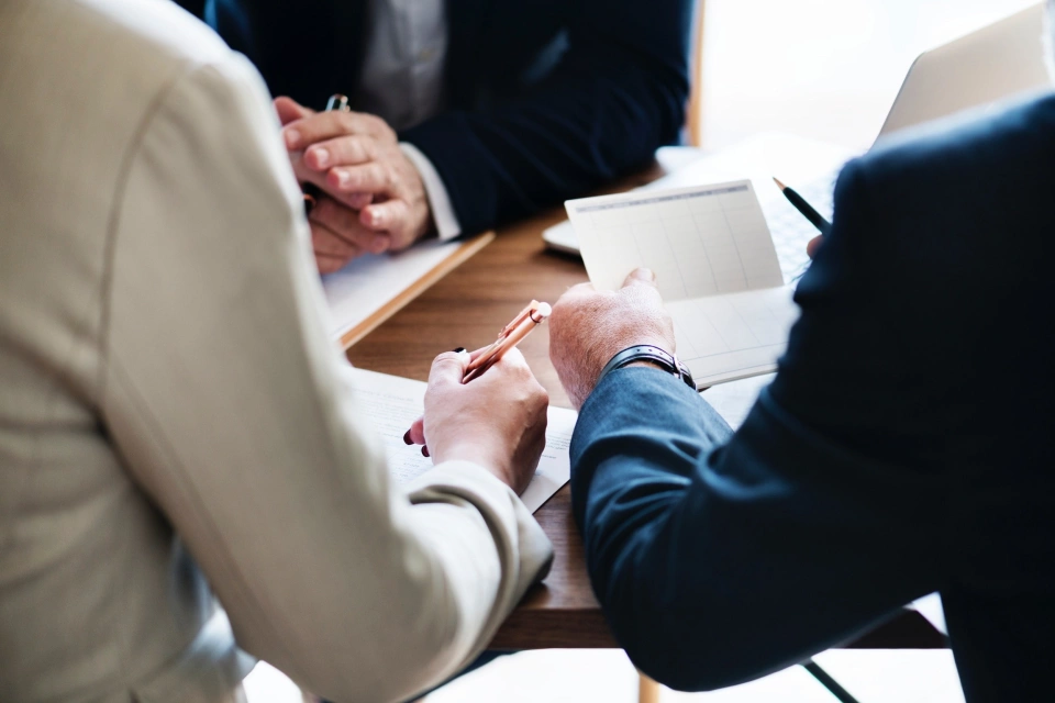 Three people sitting around a table, zoomed in on their arms and hands. One set of hands are crossed while another is holding a pen over some paperwork and the last person is holding up some documents.