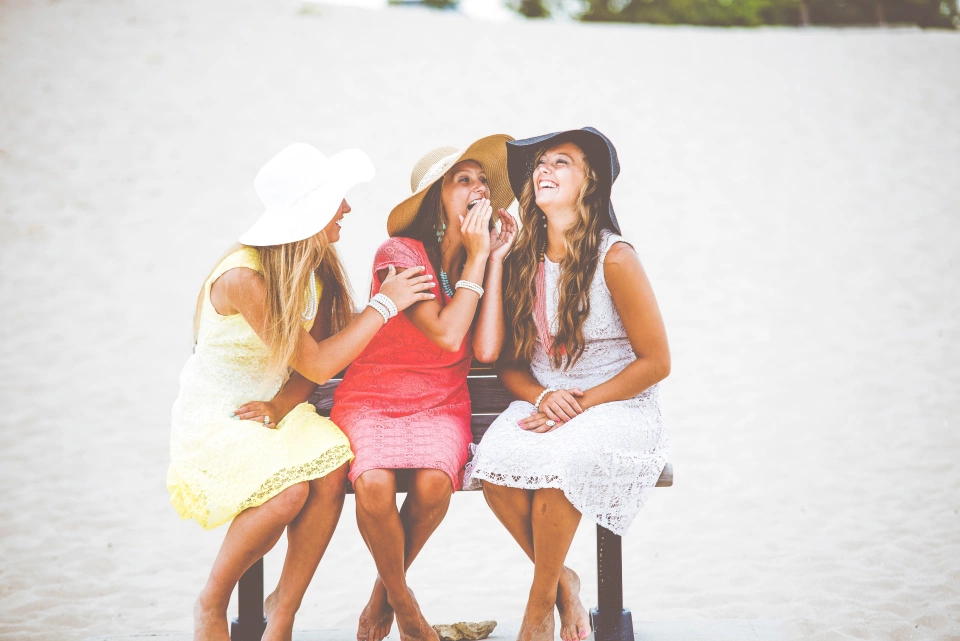 Three girls sitting on a bench at the beach with sun hats on, whispering and laughing to one another.