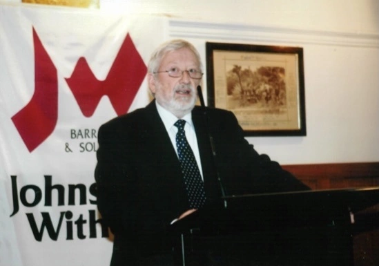 Brian Withers giving a speech behind a podium with a microphone. He is standing in front of a Johnston Withers banner and a black and white framed photo on the wall.