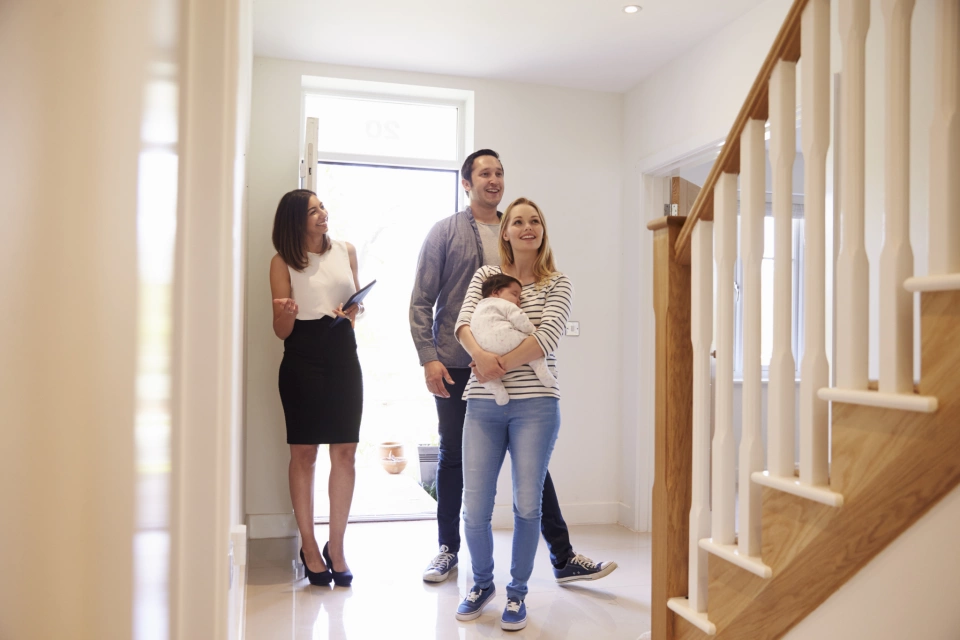 A family of three being shown around an open house with a real estate agent. The mother is holding her child while her and her husband are looking very happy at the house.