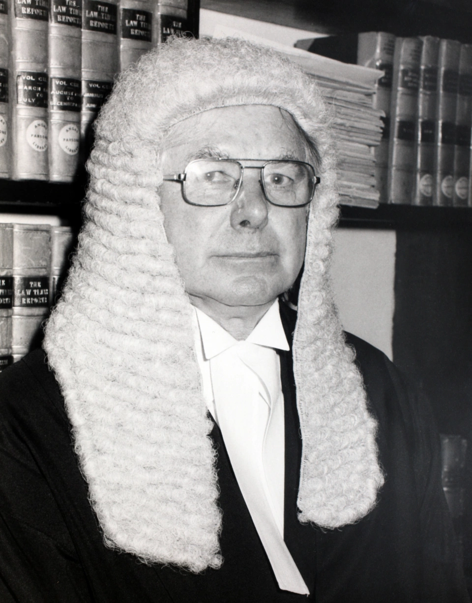 Black and White photo of Elliot Johnston dressed in a judges robe and wig in front of a bookcase of law books.